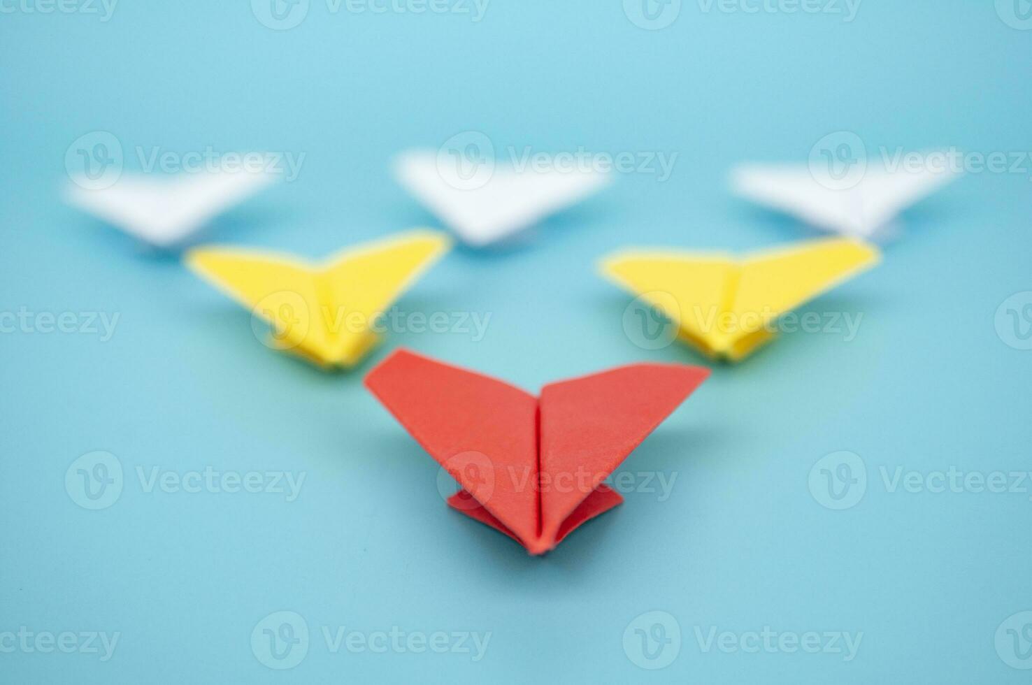 Close up view of red paper airplane origami leading yellow and white paper airplanes on blue cover background photo
