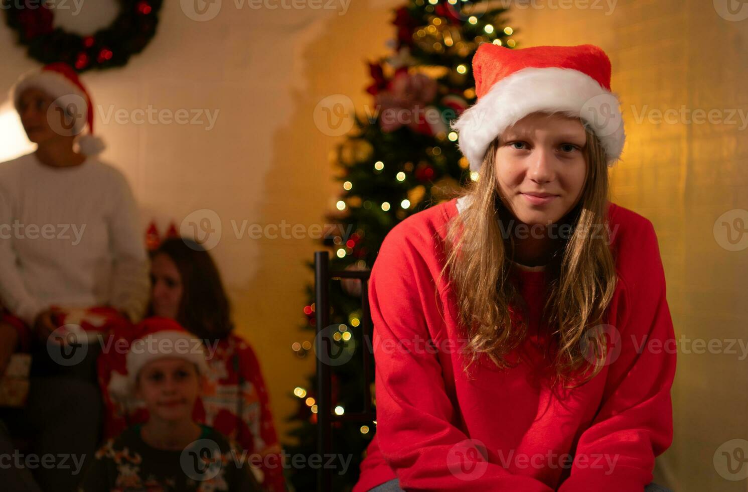 Portrait of teenage girl in Santa hat sitting on chair in front of Christmas tree, Merry Christmas and Happy Holidays photo