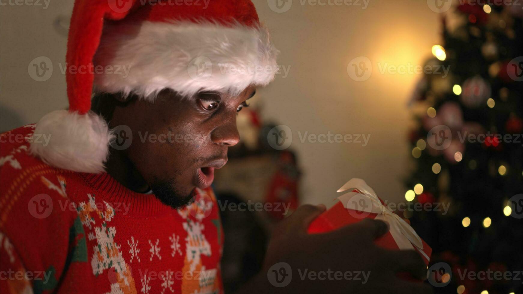 joven hombre sentado en el silla en frente de el Navidad árbol y apertura un regalo caja, alegre Navidad y contento Días festivos foto