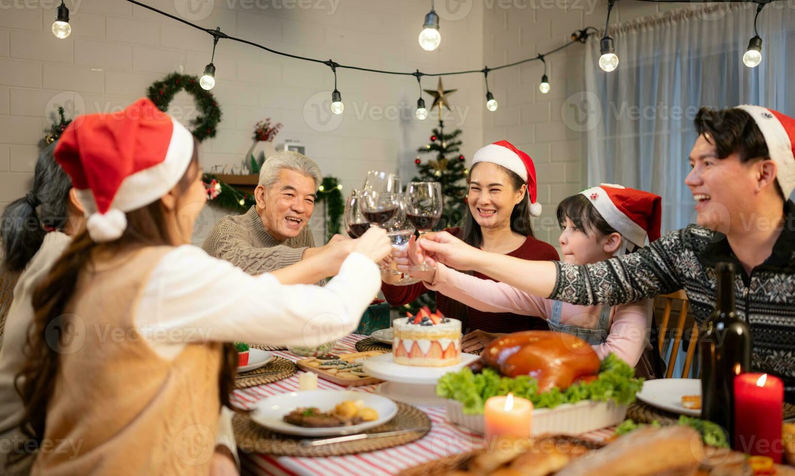 contento asiático familia celebrando Navidad juntos a hogar. alegre mayor padres y niños en Papa Noel sombrero tintinamente lentes de rojo vino. foto