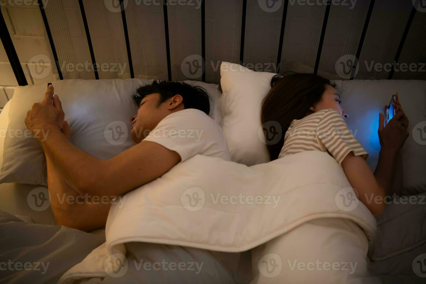 Young couple sleeping on the bed in the bedroom at night time with their backs to each other and playing on their phones, ignoring each other. photo