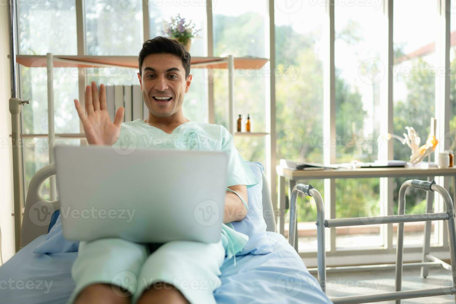 Happy young man using laptop computer while sitting on bed in hospital ward photo