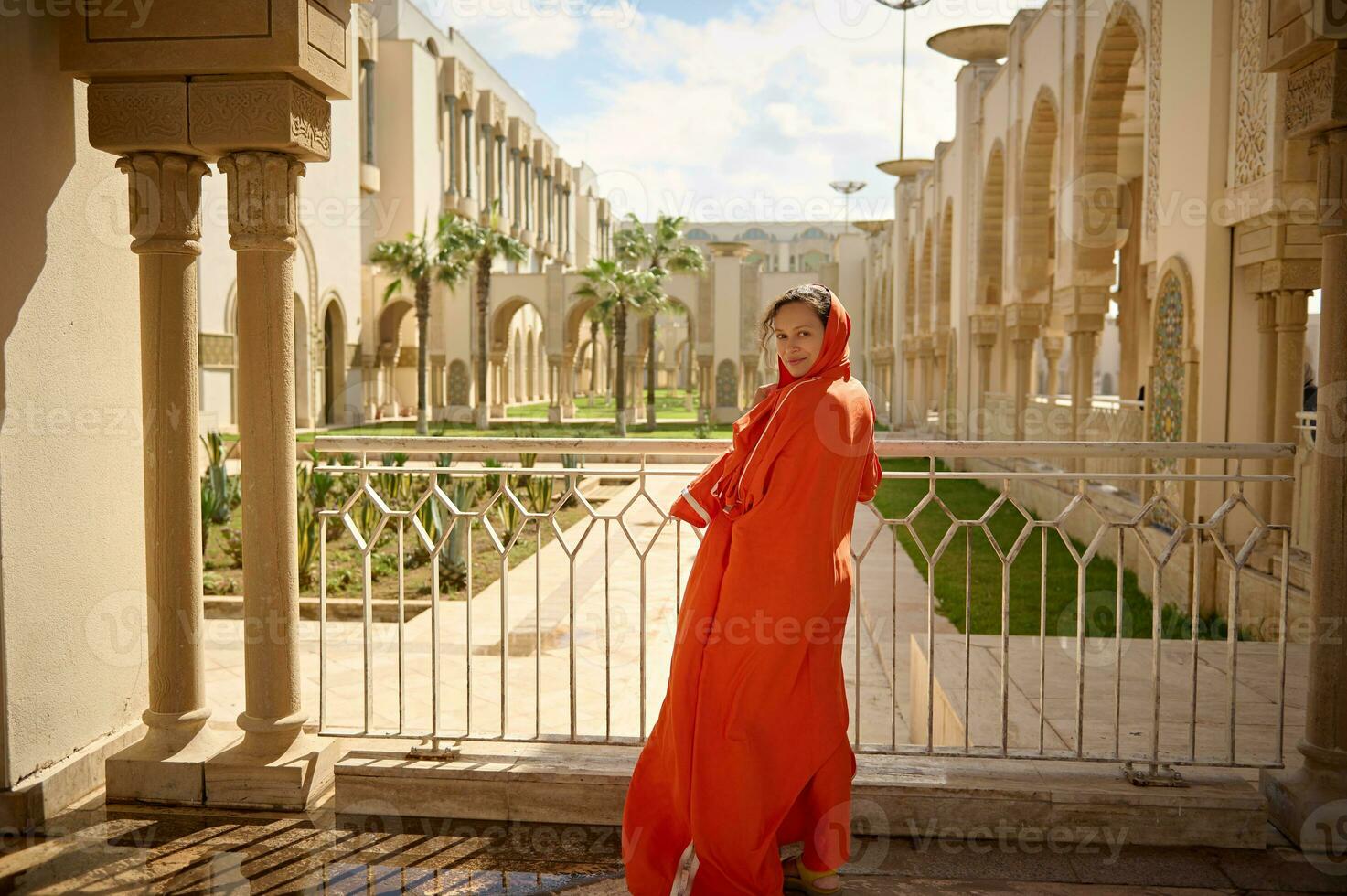 hermosa musulmán mujer con cabeza cubierto en un cabeza bufanda, a un mezquita, sonriente mirando a cámara, mediante su espalda foto