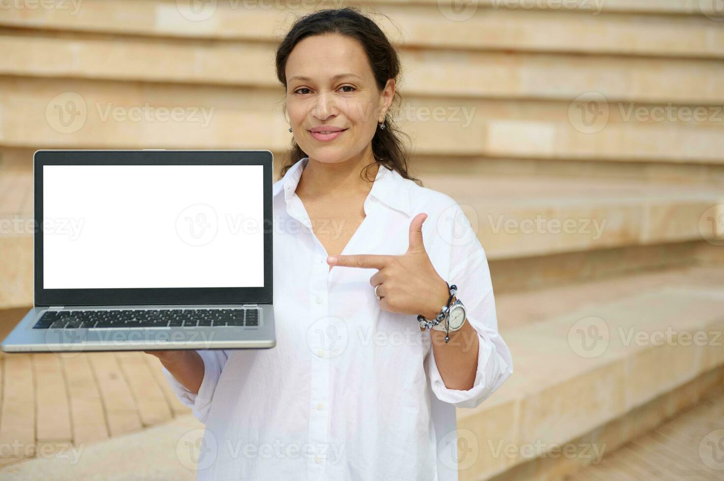 Young woman in white shirt, pointing index finger at white mockup digital screen with copy space, smiling at camera photo