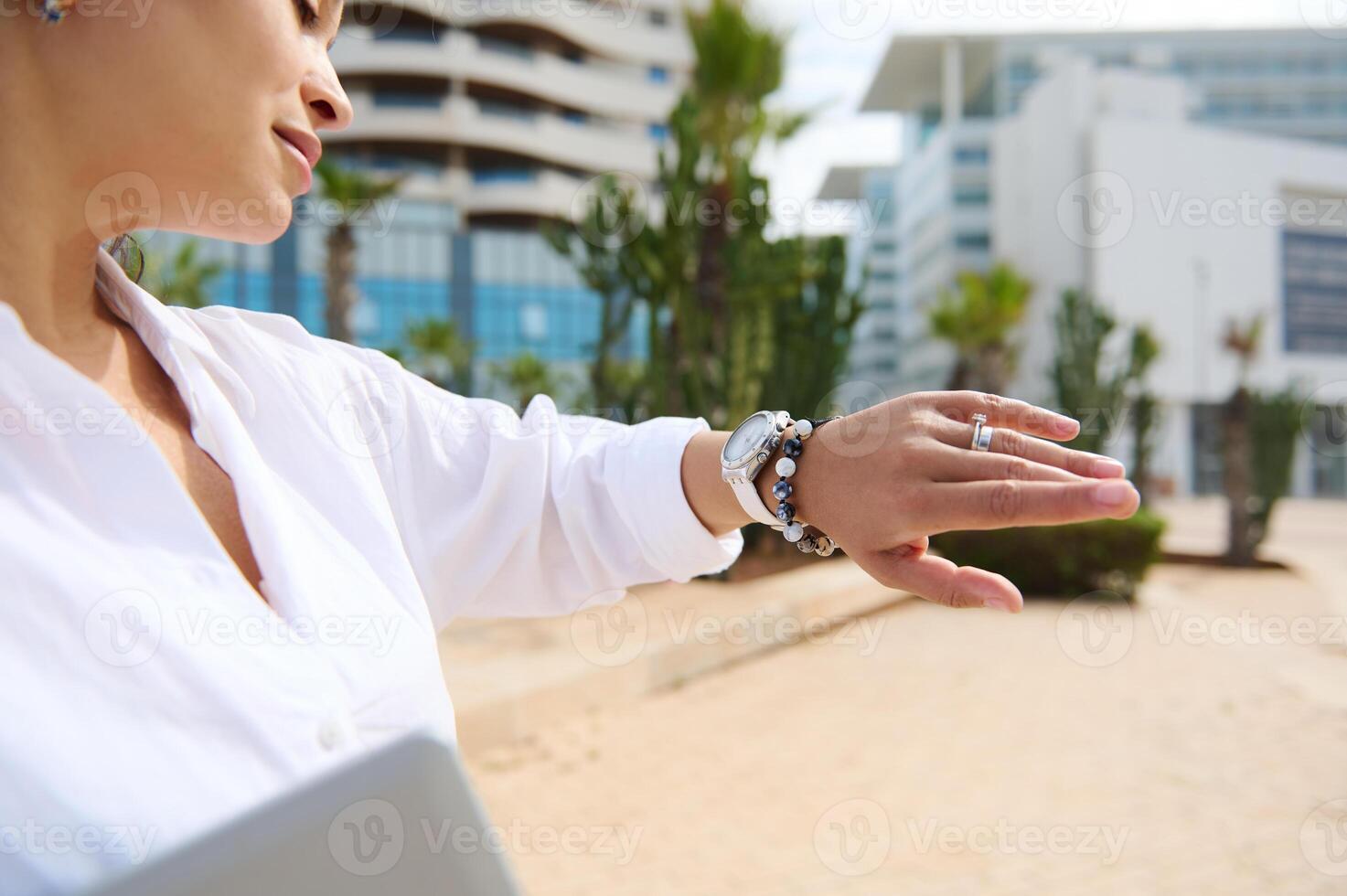 Details on the hand with stylish wrist watch of a middle aged woman checking time, standing against skyscraper backdrop photo