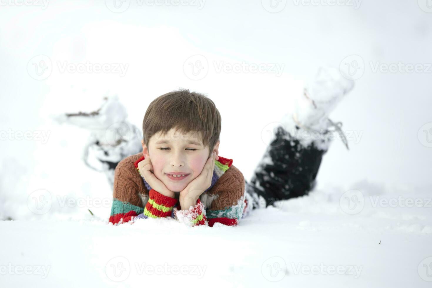 A funny little boy in colorful clothes lies in the snow. A hardened child in winter. photo