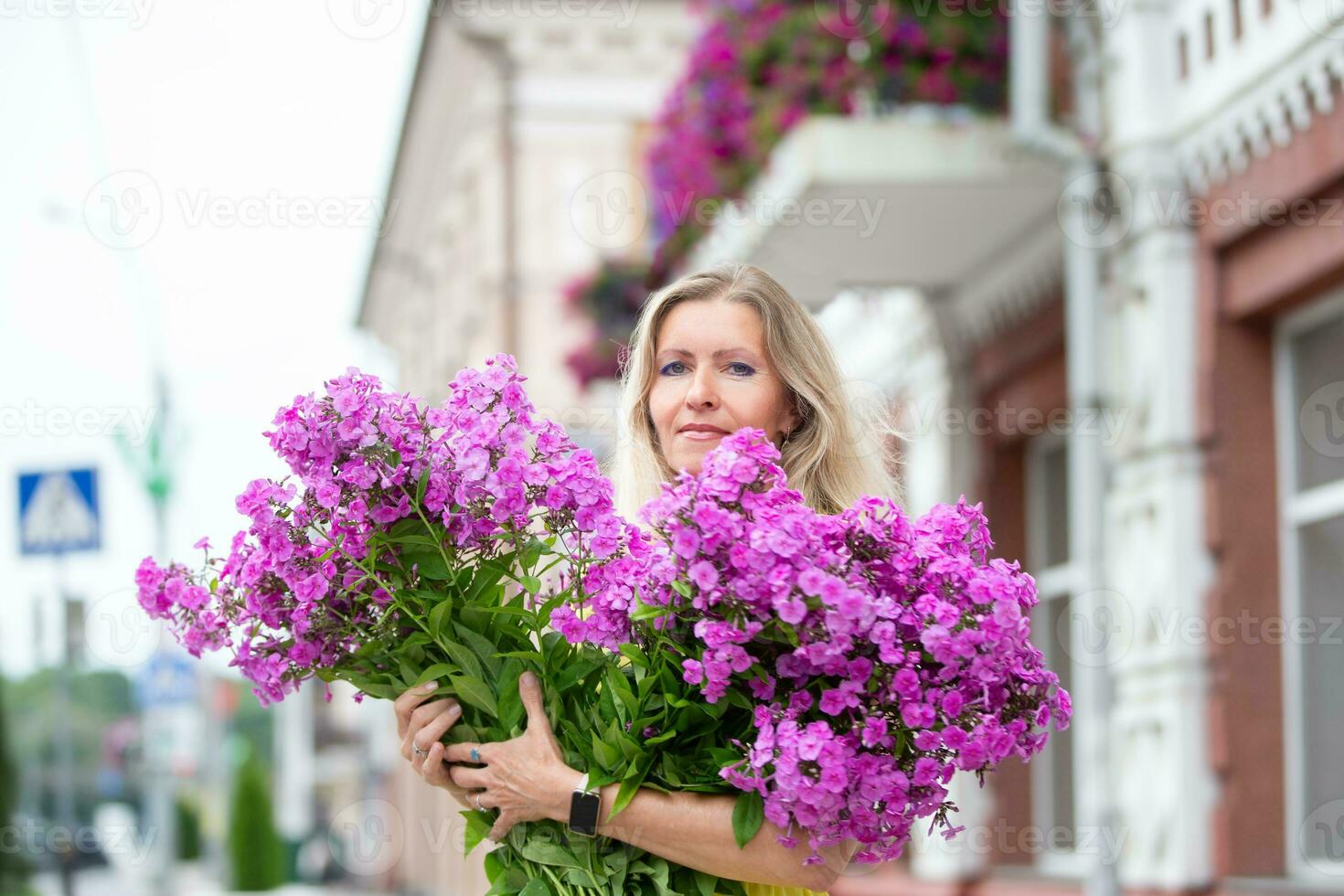 A beautiful elderly woman with a bouquet of flowers walks around the city. Age model blond with blue eyes is happy. A woman of fifty years walks in the summer, having a good time. photo