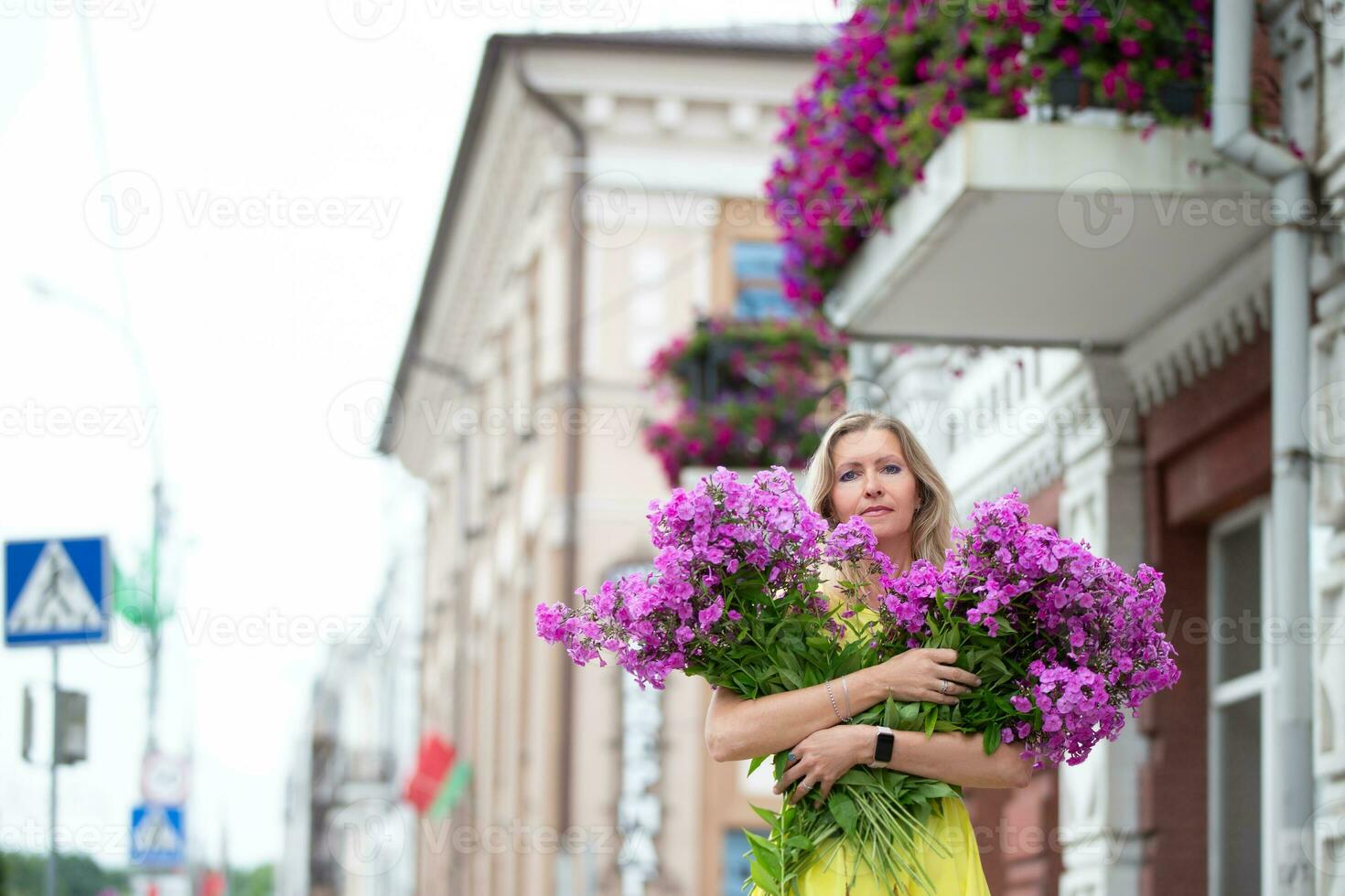 A beautiful elderly woman with a bouquet of flowers walks around the city. Age model blond with blue eyes is happy. A woman of fifty years walks in the summer, having a good time. photo