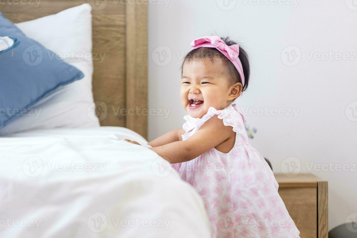 Asian baby toddler in cute pink dress is smiling while start practice walking and balancing herself at the bed with happiness for healthy kid and adorable girl portrait usage photo