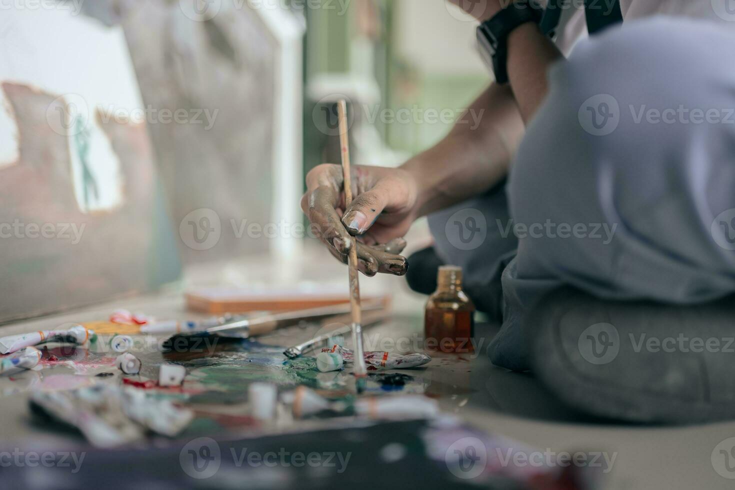 Selective focus. Oil painting techniques. Close up view of male artist holding brush mixing oil paints in colorful plastic palette on floor. photo