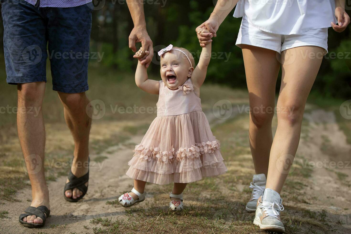 Happy one-year-old girl is being held by dad and mom. Little child walking with parents. A cute baby learns to walk with the help of her parents. photo