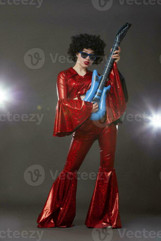 A full-length girl in a flared disco-style stage costume holds a comic guitar in her hands. photo