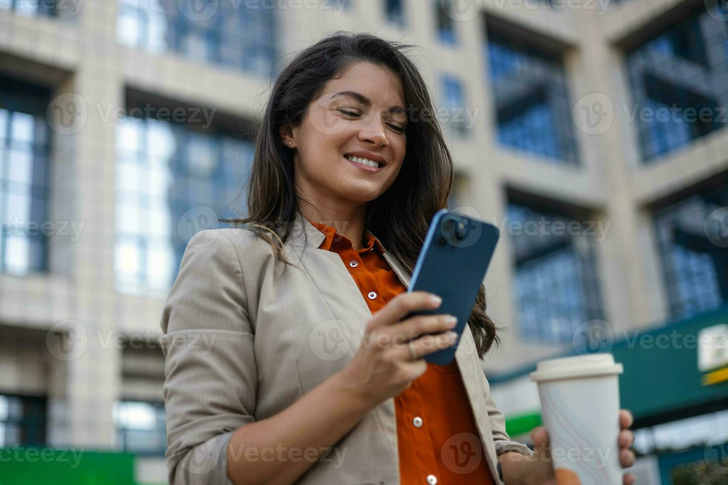 Young smiling brunette woman holding a smartphone and a cup of coffee looking at device screen against the background of an office building. copy space photo