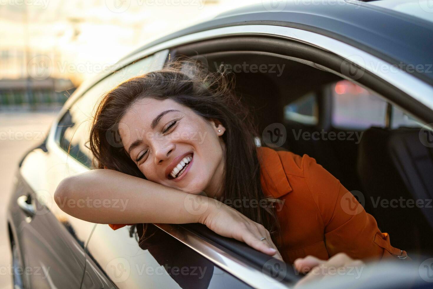 alegre joven hembra sentado en brillante coche en pasajero asiento y propensión fuera abierto ventana mientras disfrutando el paseo foto