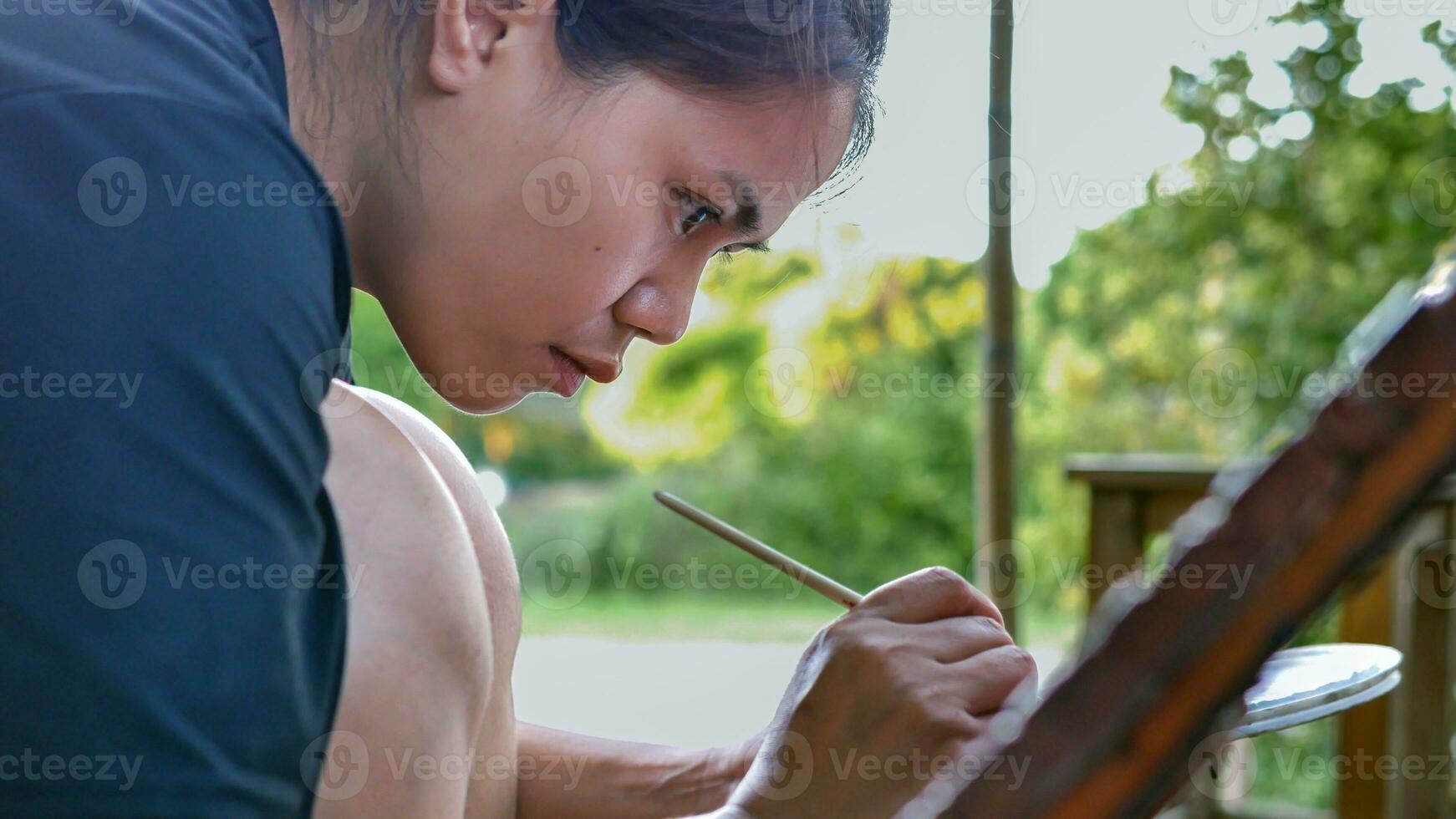 Close-up of Asian woman painter creating art use a paintbrush to draw lettering designs on a wooden coffee shop sign. outdoor activities, People doing activities. photo