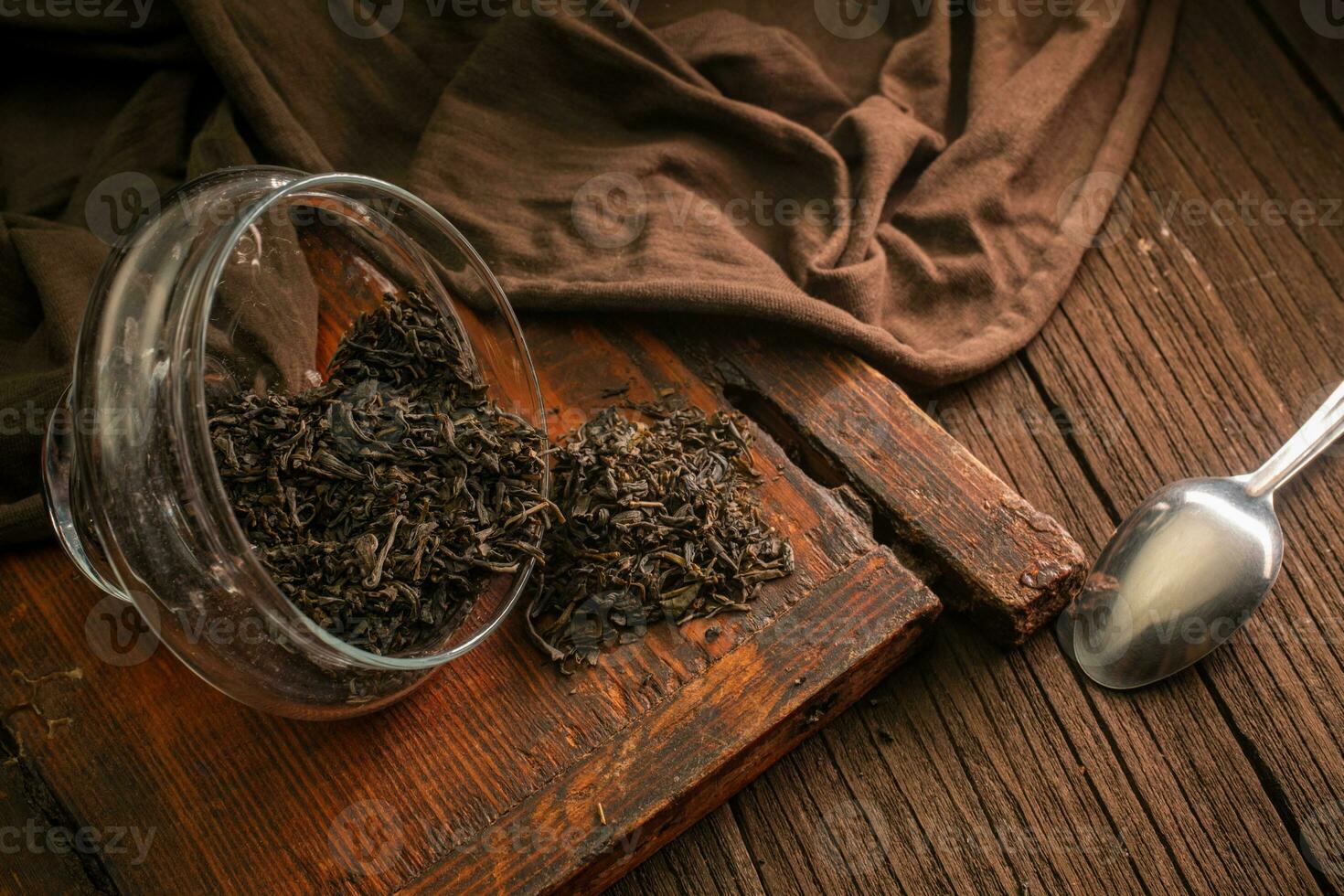 Dried leaf black tea is scattered from glass jars on a wooden vintage board with a spoon and a cloth on a dark background photo