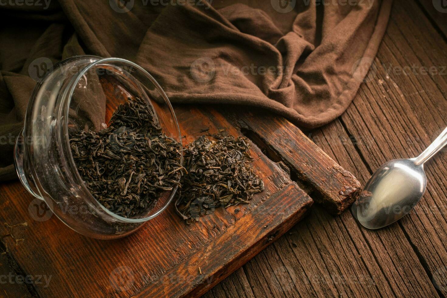 seco hoja negro té es dispersado desde vaso frascos en un de madera Clásico tablero con un cuchara y un paño en un oscuro antecedentes foto