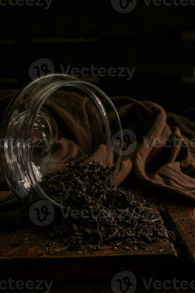 Black delicious leaf dried tea poured out of a glass jar on a wooden vintage brown table on a dark background photo