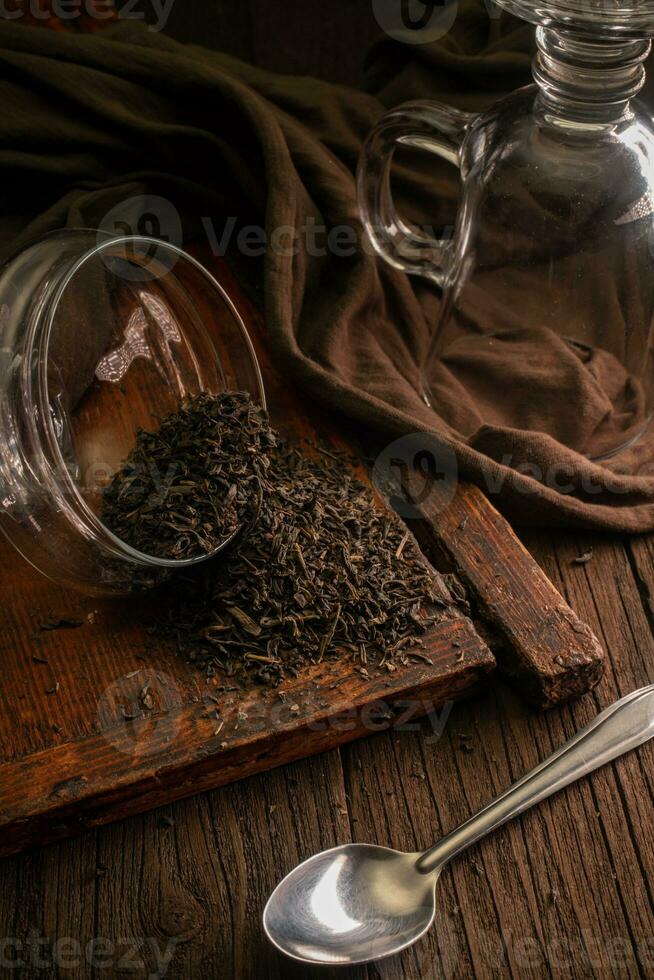 Dried leaf black tea is scattered from a glass jar on a wooden brown board with a spoon, a glass and a cloth on a dark background photo