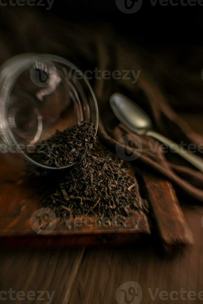 delicioso negro seco suelto hoja té es dispersado desde vaso frascos en un de madera Clásico mesa en un cuchara y un paño en un oscuro antecedentes con un bokeh efecto foto