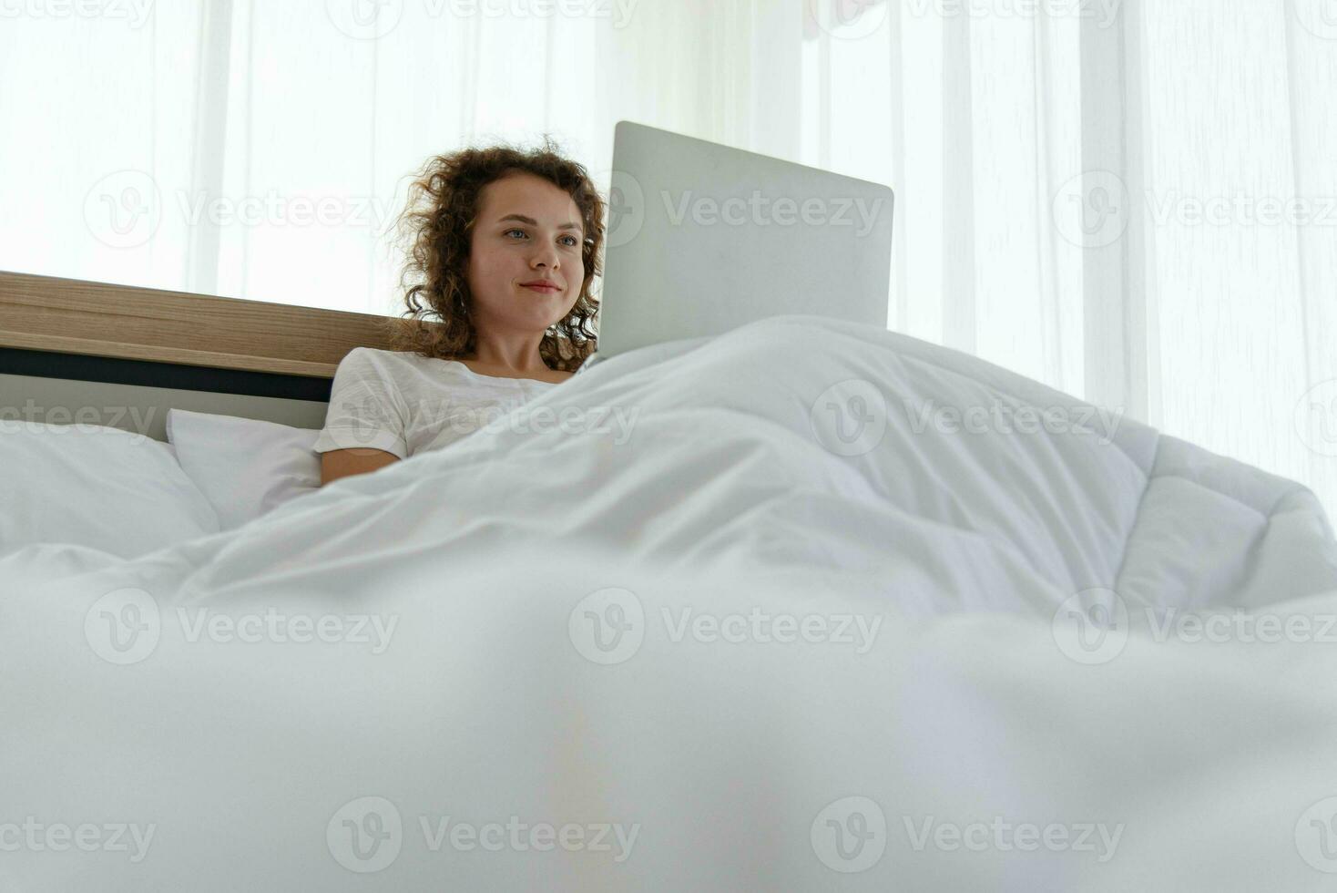 Young beautiful happy woman relaxing and working with laptop while lying in bed in the morning after waking up. photo