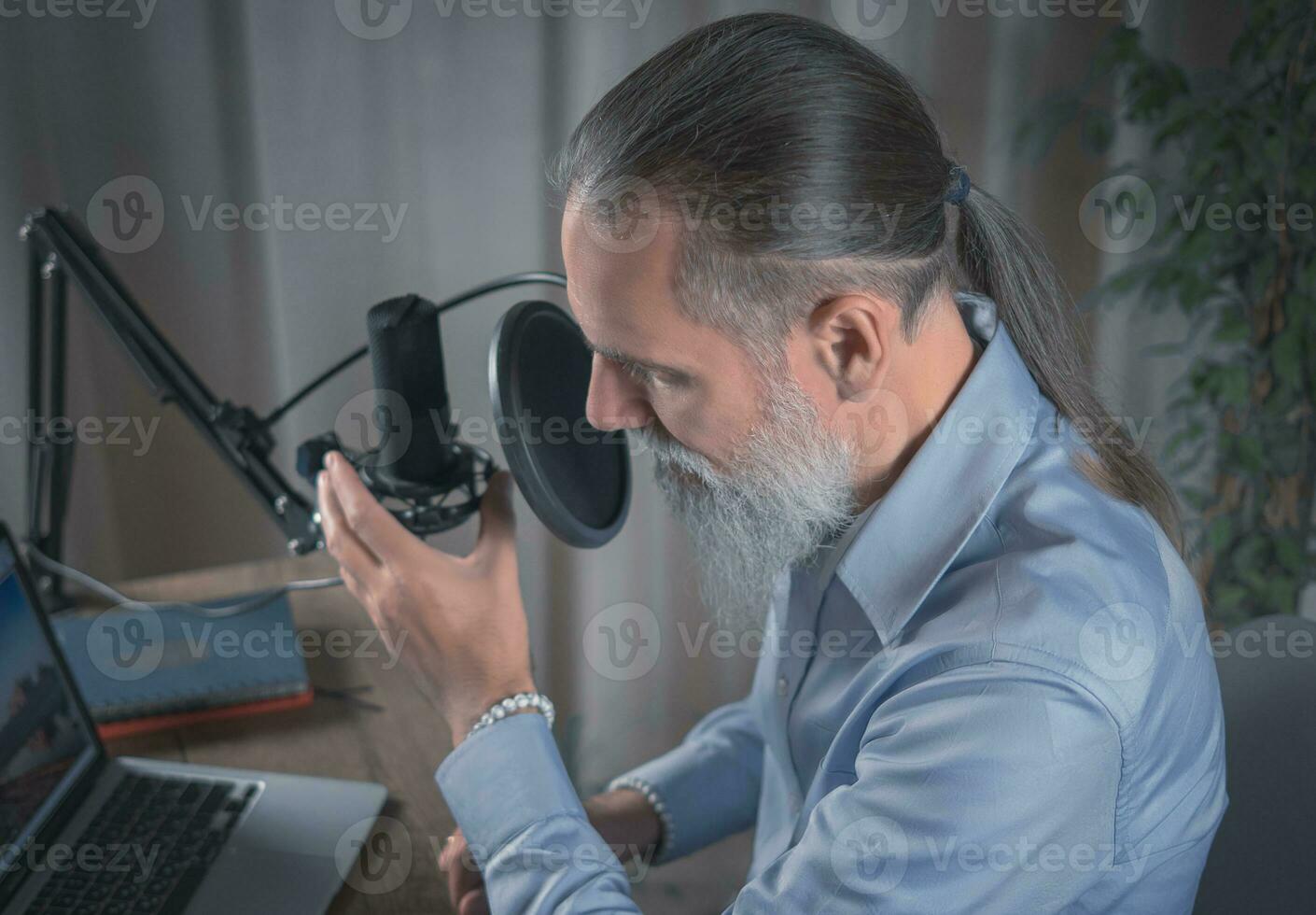 A man with a beard wearing glasses broadcasts his audio podcast using a microphone and laptop in a home studio. Close-up photo