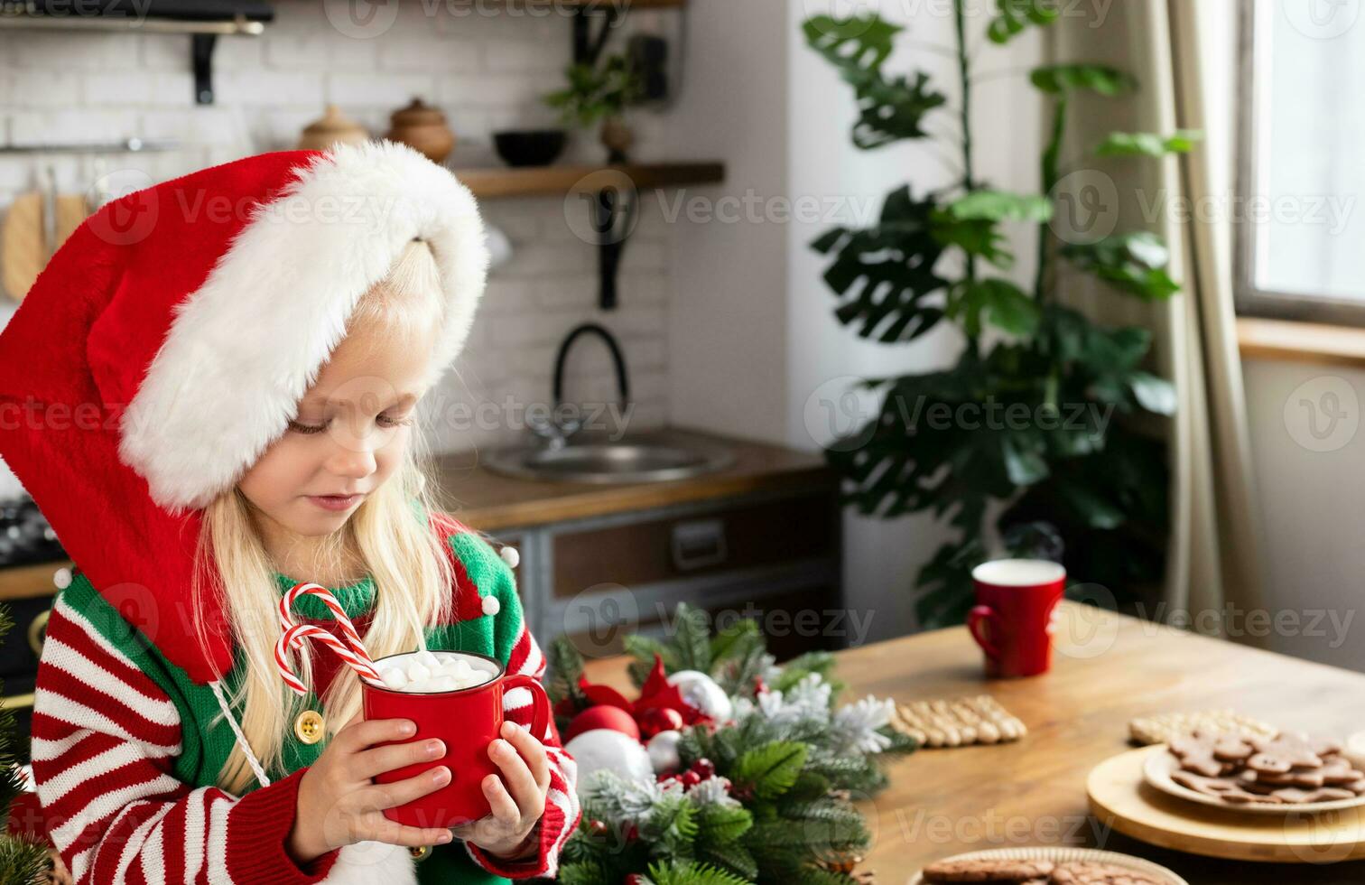 Kid sitting on Christmas decorated counter. Child serious and looking at red mug with beverage and candy canes in photo