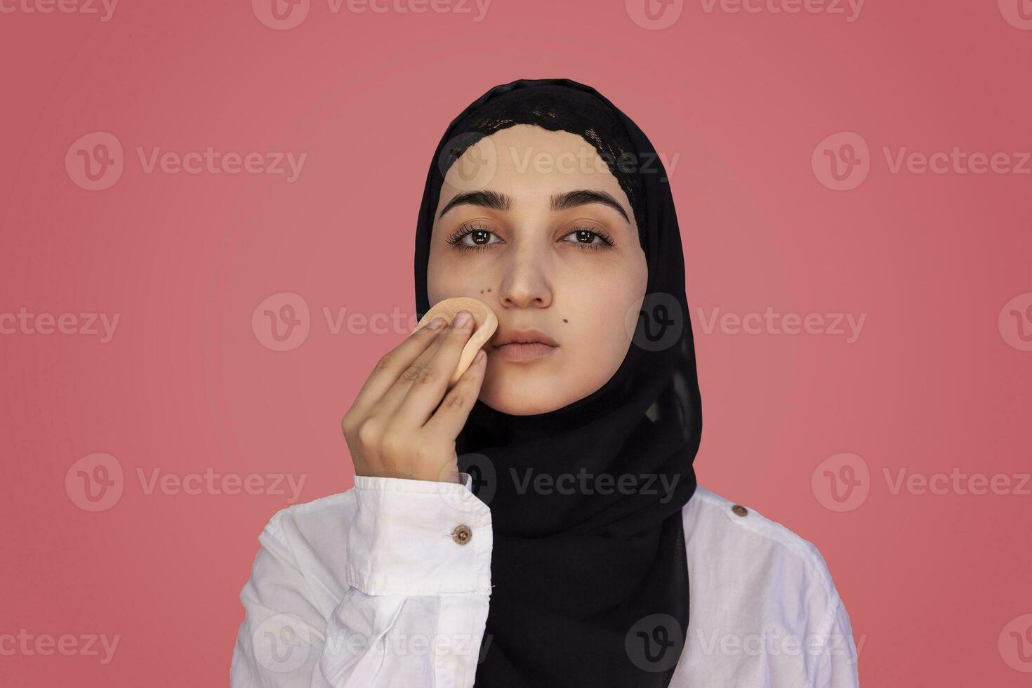 Cute 20s female applying applying powder with sponge on her face. Beautiful Persian woman in hijab doing make-up photo