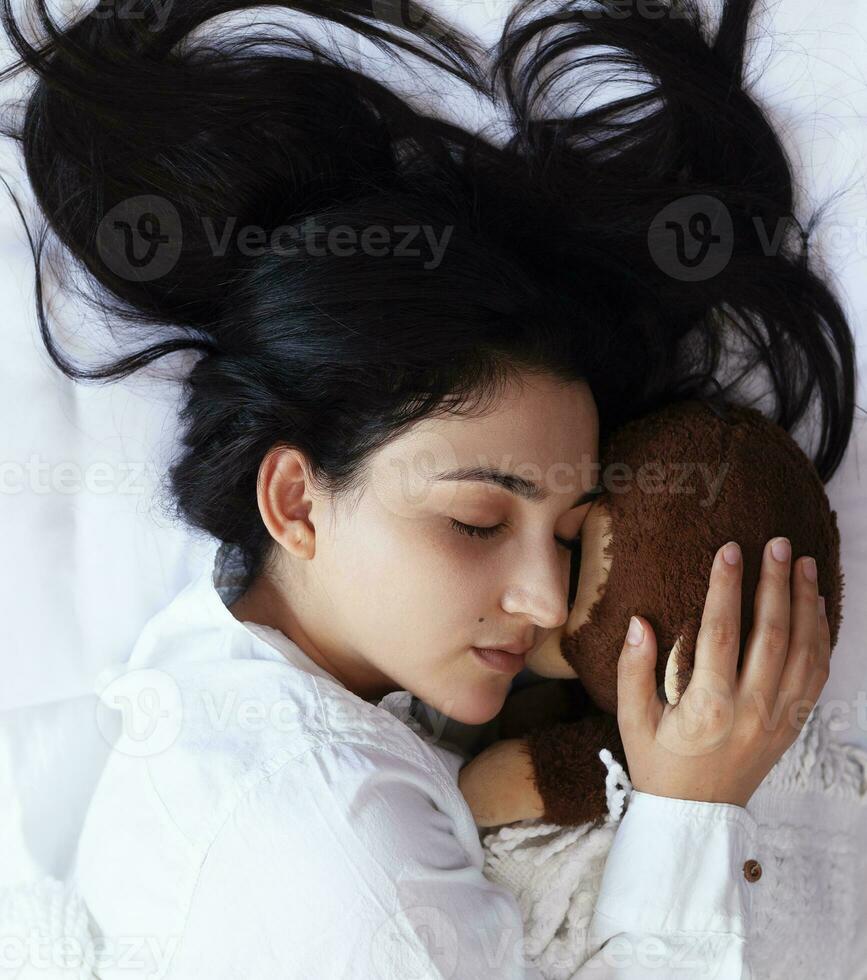 Young woman sleeping with toy bear. Cute black haired woman resting in bed with pleasure. Happy life of single lady. Pretty girl enjoying long sleep on her weekend photo