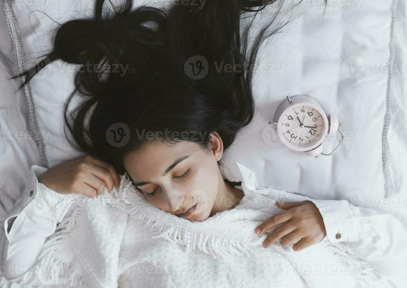 Beautiful black-haired woman sleeping in cozy bed. The alarm clock set at 7 a.m. morning. Female is going to wake up at 7 o'clock early morning. Top view photo