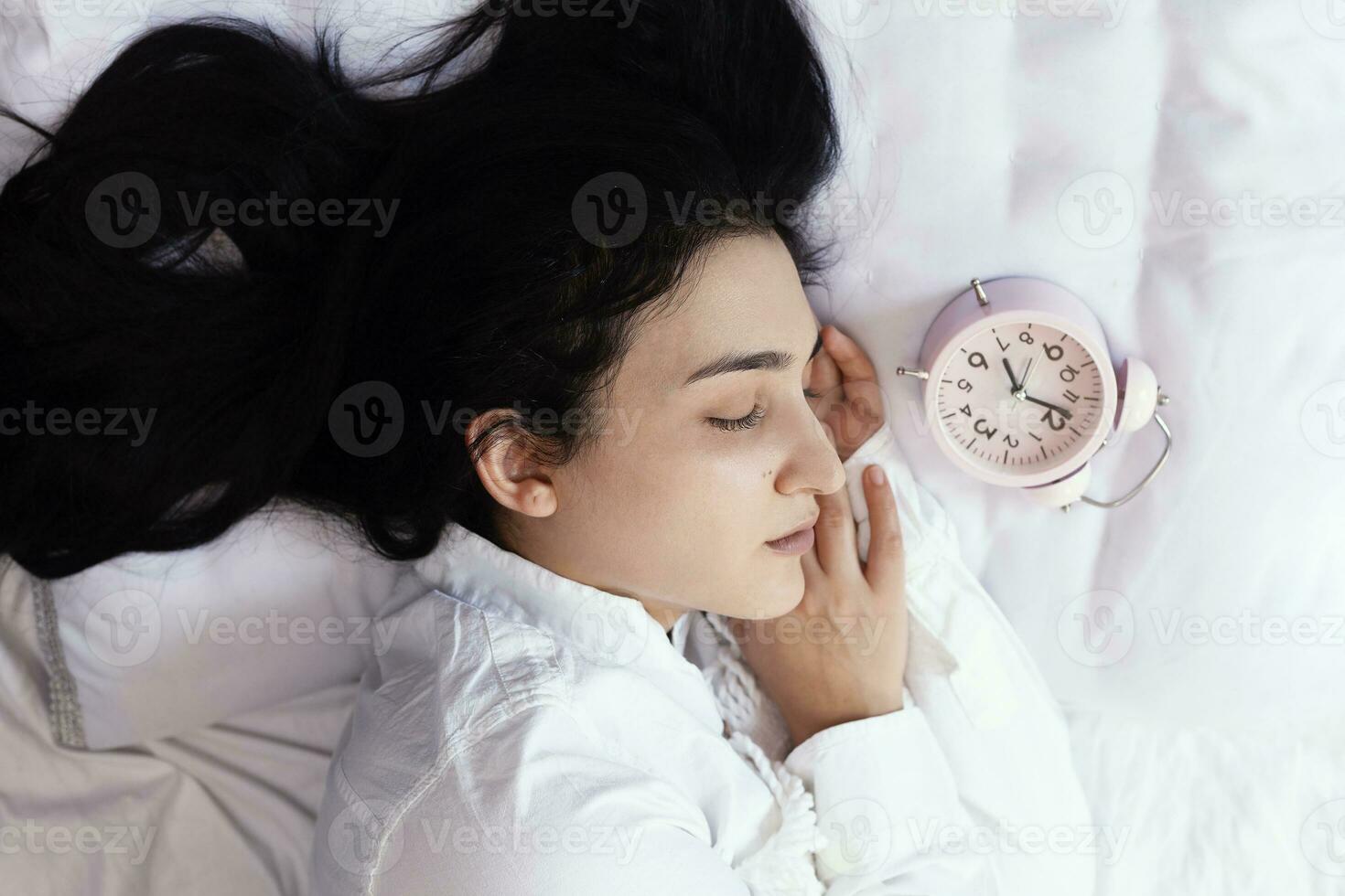 Beautiful black-haired woman sleeping in cozy bed. The alarm clock set at 7 a.m. morning. Female is going to wake up at 7 o'clock early morning. Top view photo