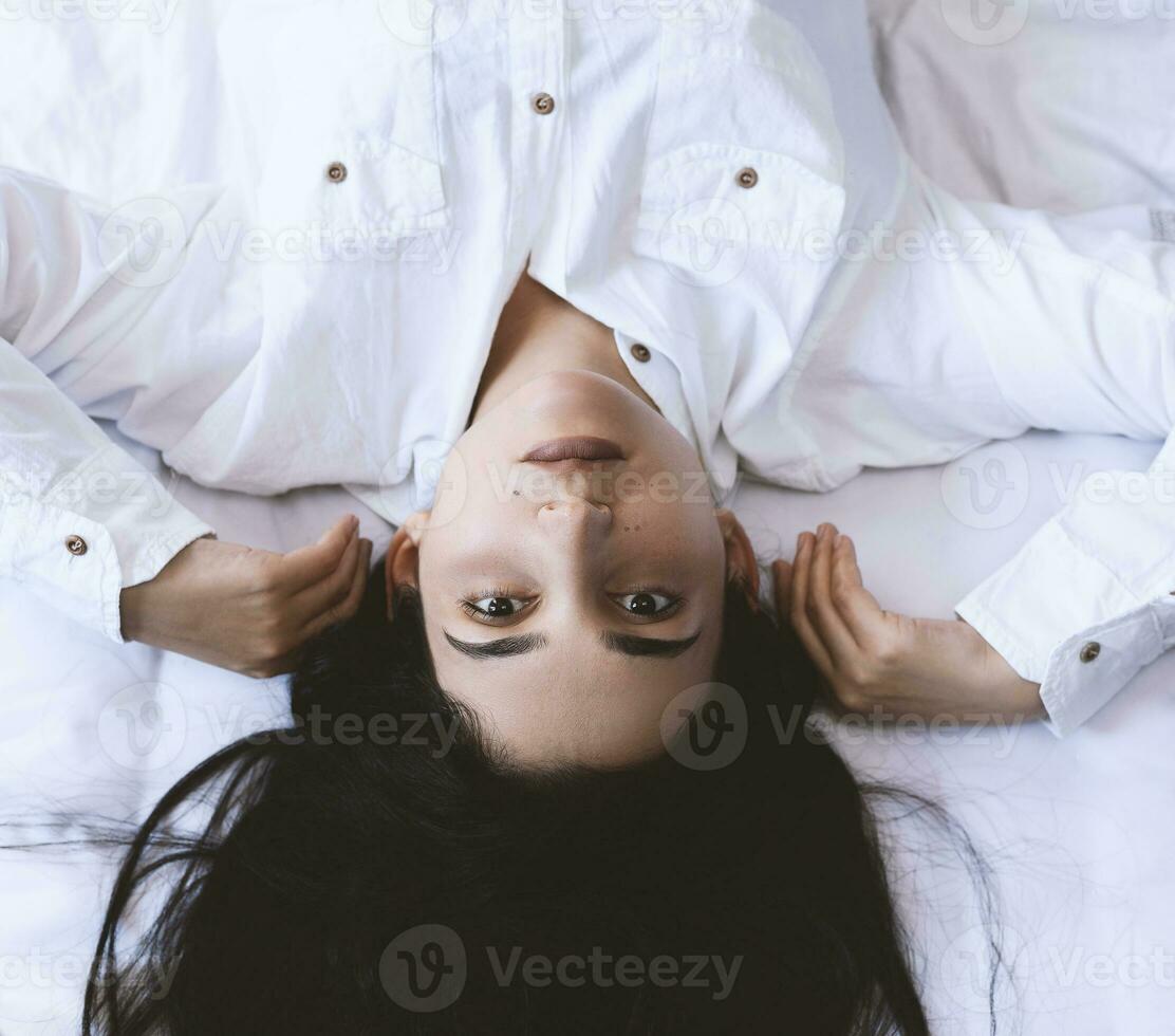 hermosa mujer acostado en cama. bonito mujer con largo morena pelos acostado en cama.alto ángulo ver de un joven contento niña vestido en un blanco camisa mentiras en un cama a hogar foto