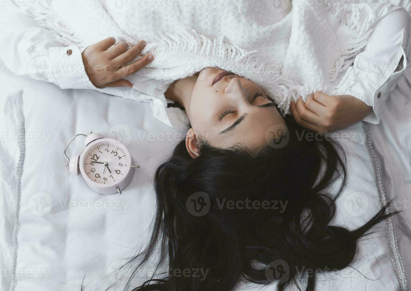 Beautiful black-haired woman sleeping in cozy bed. The alarm clock set at 7 a.m. morning. Female is going to wake up at 7 o'clock early morning. Top view photo