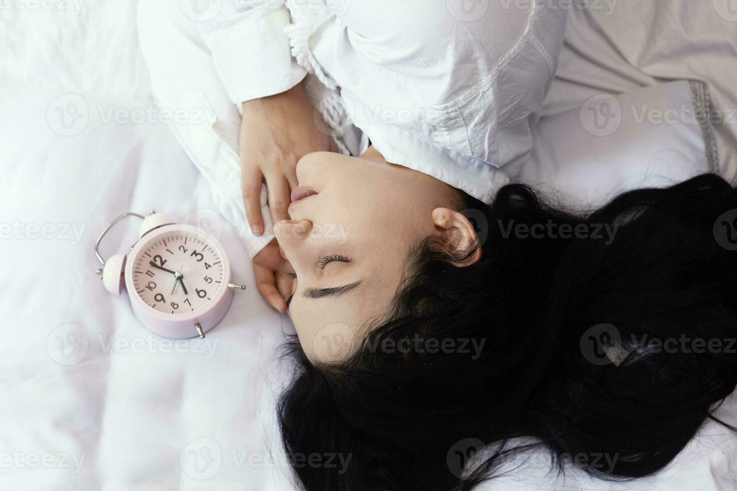 Beautiful black-haired woman sleeping in cozy bed. The alarm clock set at 7 a.m. morning. Female is going to wake up at 7 o'clock early morning. Top view photo