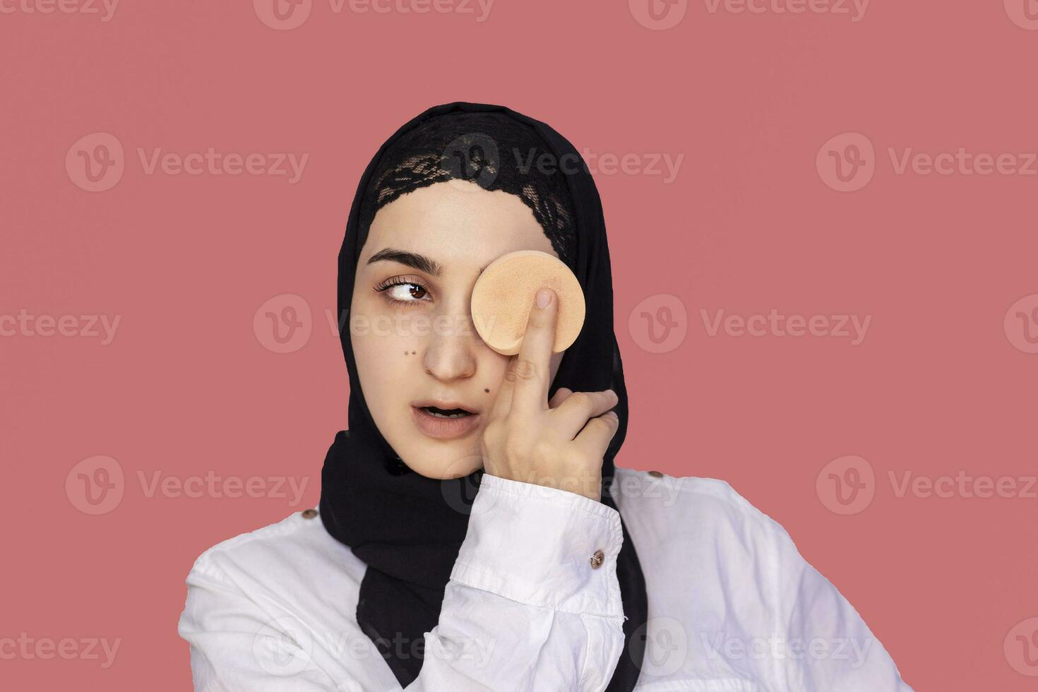 Cute 20s female applying applying powder with sponge on her face. Beautiful Persian woman in hijab doing make-up photo