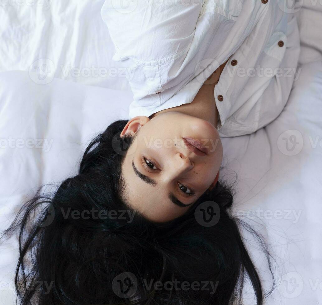 Beautiful woman lying in bed. Pretty woman with long brunette hairs lying in bed.High-angle view of a young happy girl dressed in a white shirt lies on a bed at home photo