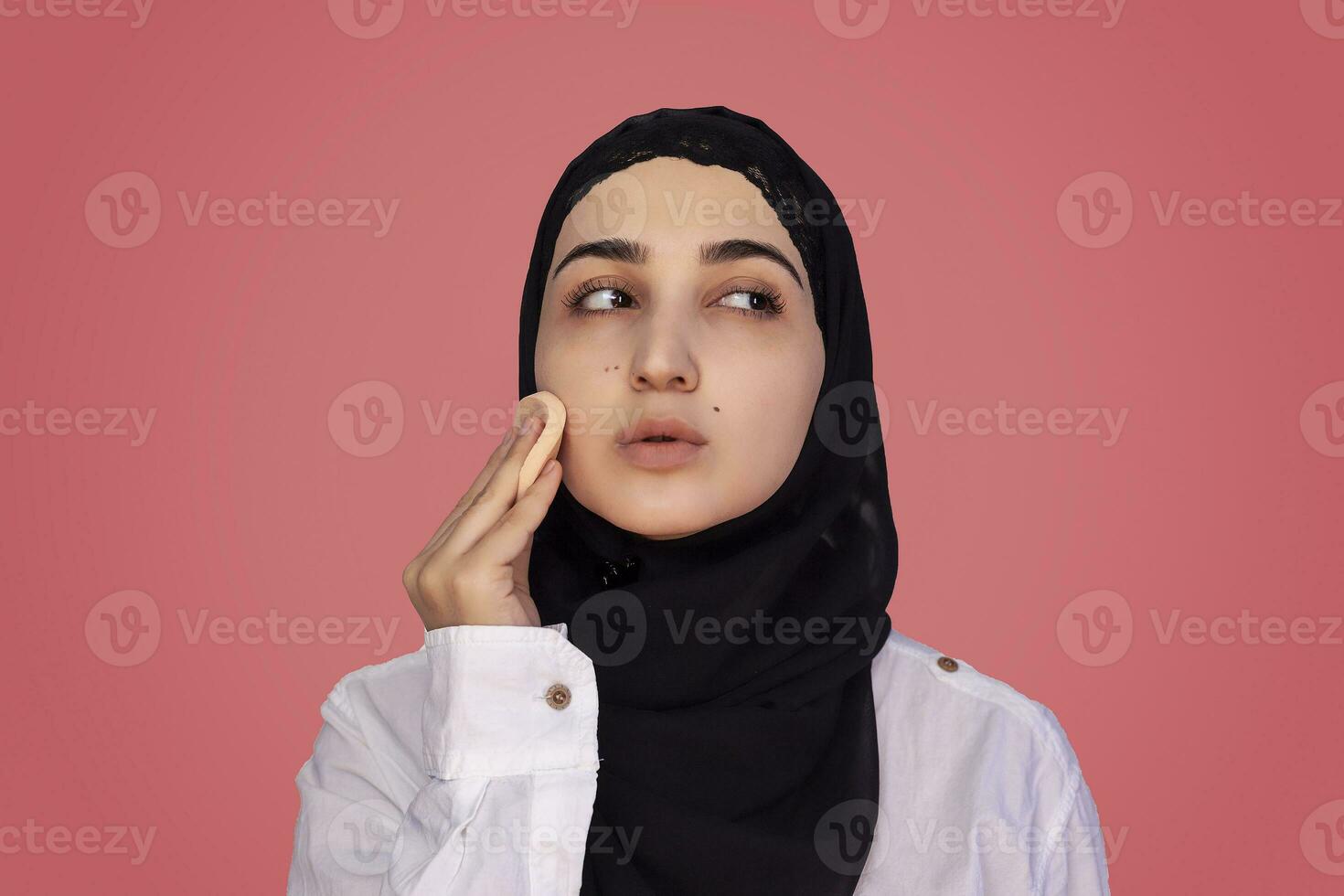 Cute 20s female applying applying powder with sponge on her face. Beautiful Persian woman in hijab doing make-up photo
