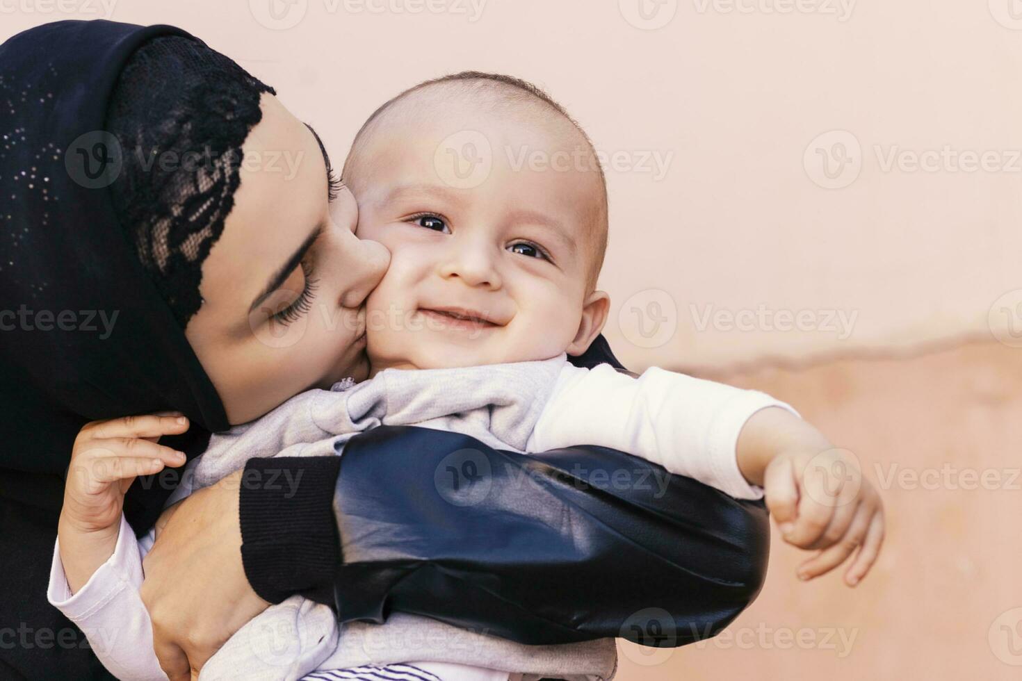 Young Muslim woman in hijab holding her 1-year-old baby boy, hugging and kissing. Happy Muslim mother in abaya clothing kiss and play with her little child photo