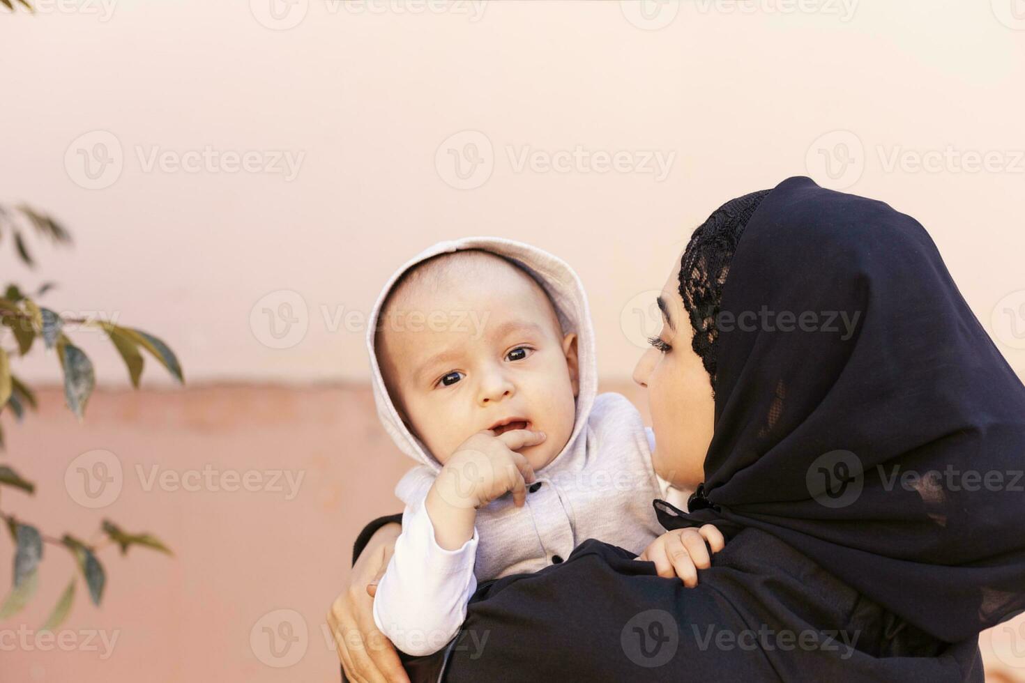 joven musulmán mujer en hijab participación su 1 año bebé chico, abrazando y besando contento musulmán madre en abaya ropa Beso y jugar con su pequeño niño foto