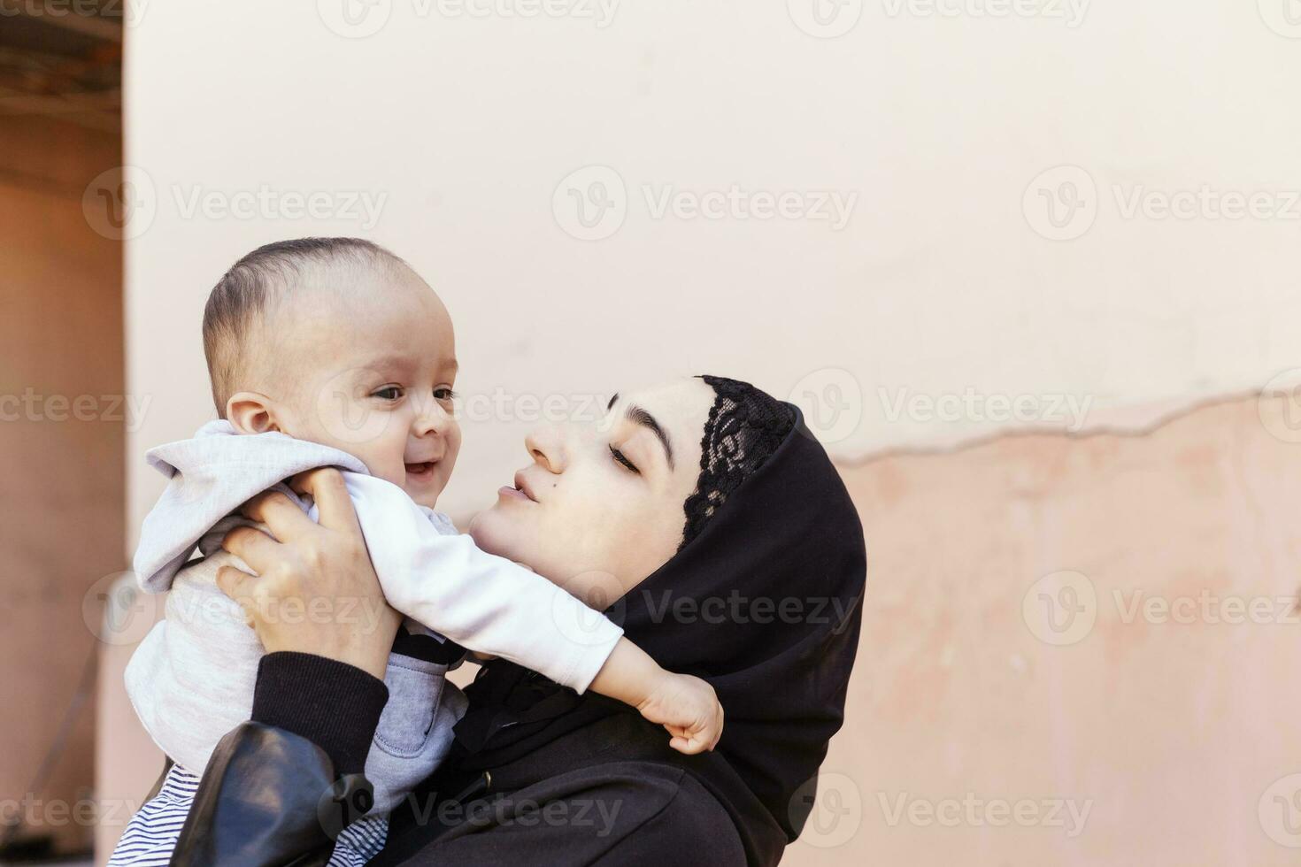 joven musulmán mujer en hijab participación su 1 año bebé chico, abrazando y besando contento musulmán madre en abaya ropa Beso y jugar con su pequeño niño foto
