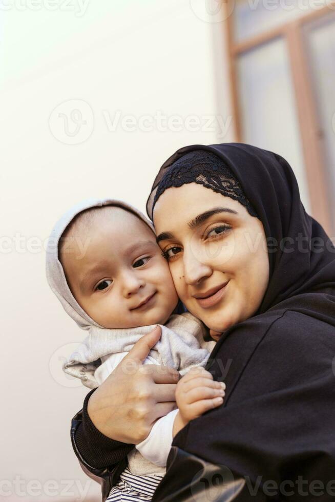 joven musulmán mujer en hijab participación su 1 año bebé chico, abrazando y besando contento musulmán madre en abaya ropa Beso y jugar con su pequeño niño foto