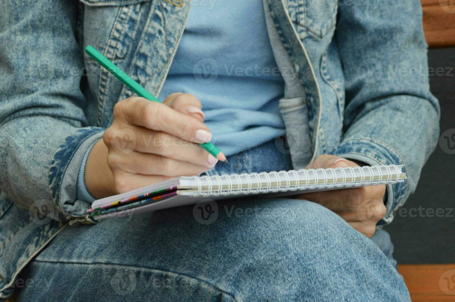 A girl, a woman in jeans and holding a notebook and a pencil, makes notes on the pages of a sketchbook, holding it on her lap, sitting on a street bench. photo