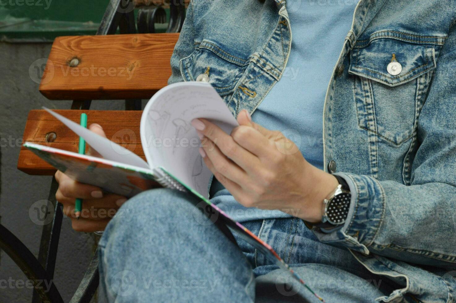 un chica, un mujer en pantalones y un chaqueta, voltea mediante el paginas de un bloc, bloc de dibujo, participación eso en su rodillas con un lápiz en su mano, hace notas, bocetos en un calle banco. foto