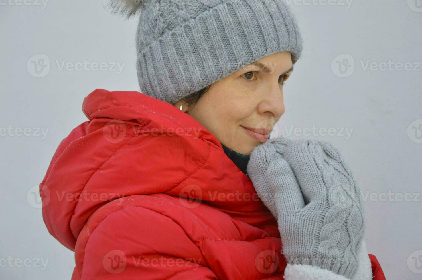 A young woman in a gray knitted hat and warm mittens holds her hands near her face. Portrait of a Caucasian woman in a red jacket with a hood on a light background. photo