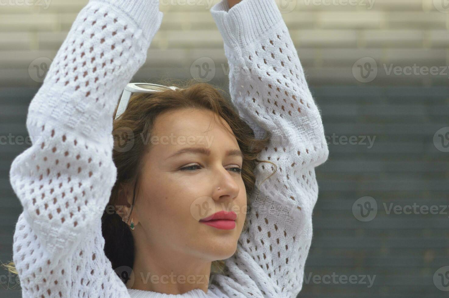 Close-up portrait of an attractive girl in a white sweater, with her hands raised up against a gray brick wall. photo