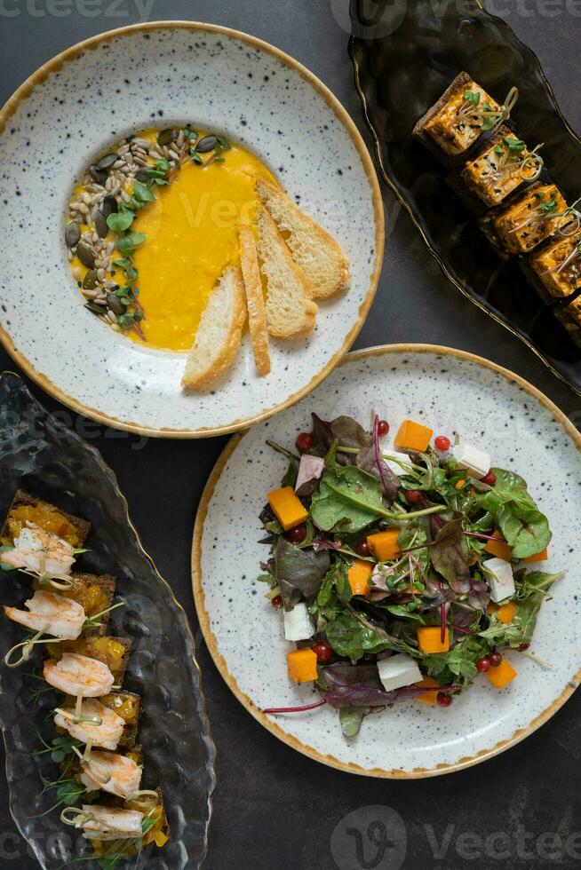 Pumpkin salad and soup, shrimp and marinated cheese canapes on dark background top view, flat lay photo