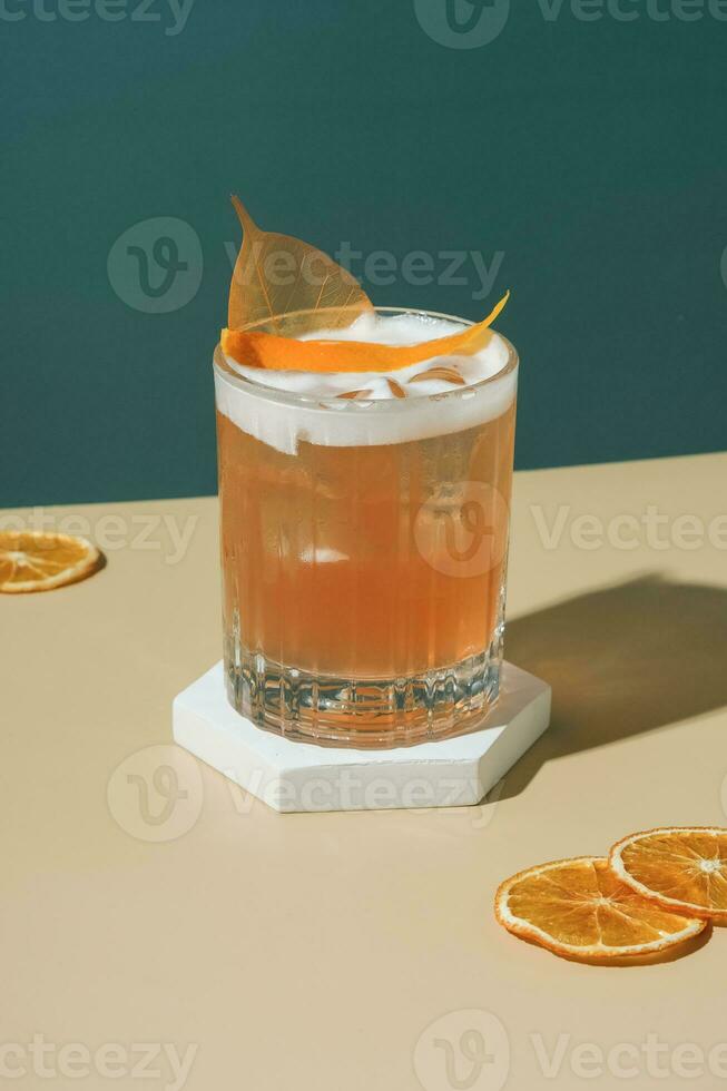 Alcoholic cocktail with ice and foam on podium with dried orange slices on coloured background. photo