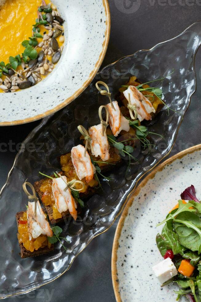 Shrimp and mango chutney canapes, pumpkin soup in glass plate on dark background top view, flat lay photo