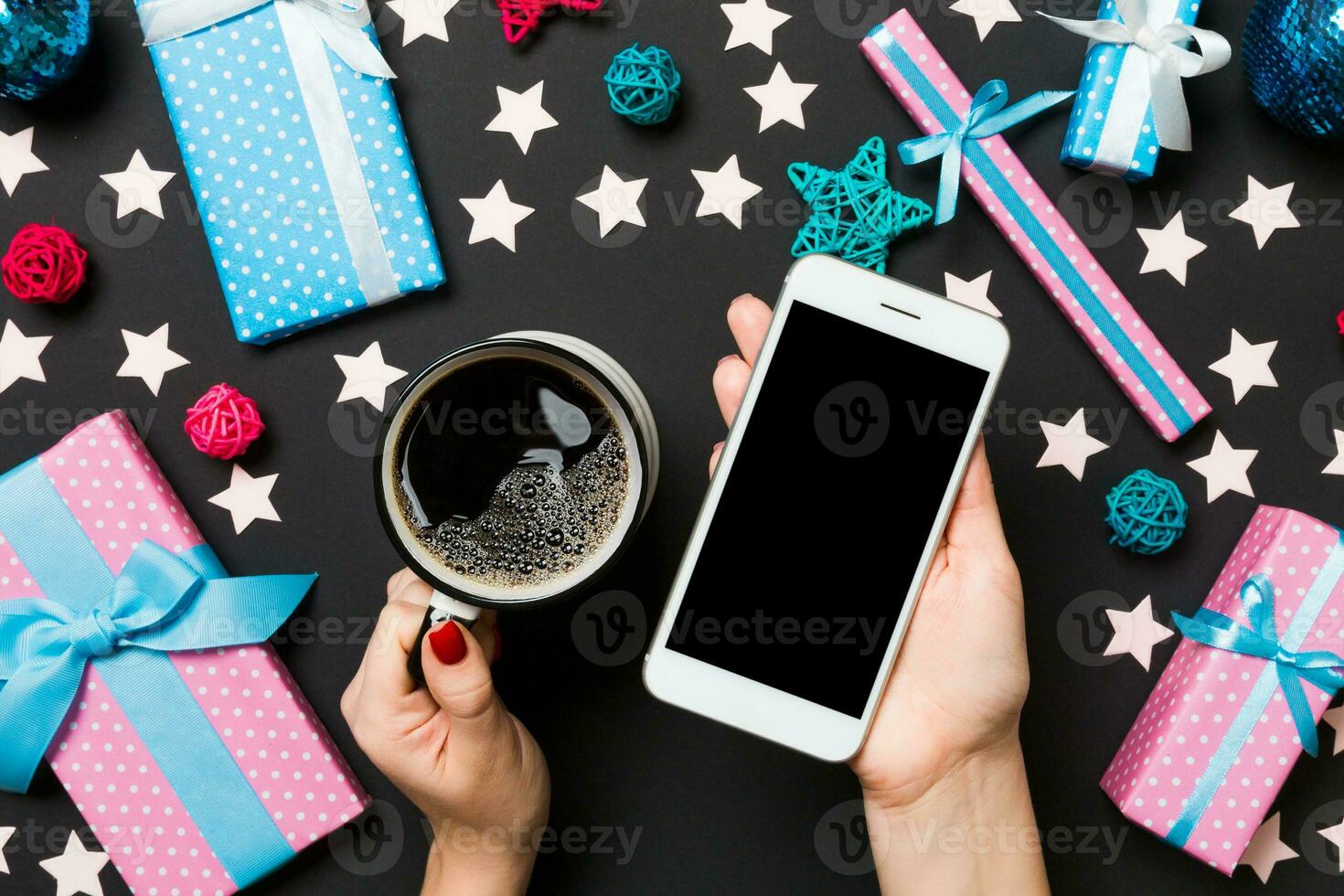 Top view of a woman holding a phone in one hand and a cup of coffee in another hand on black background. Christmas decorations and toys. New Year holiday concept. Mockup photo