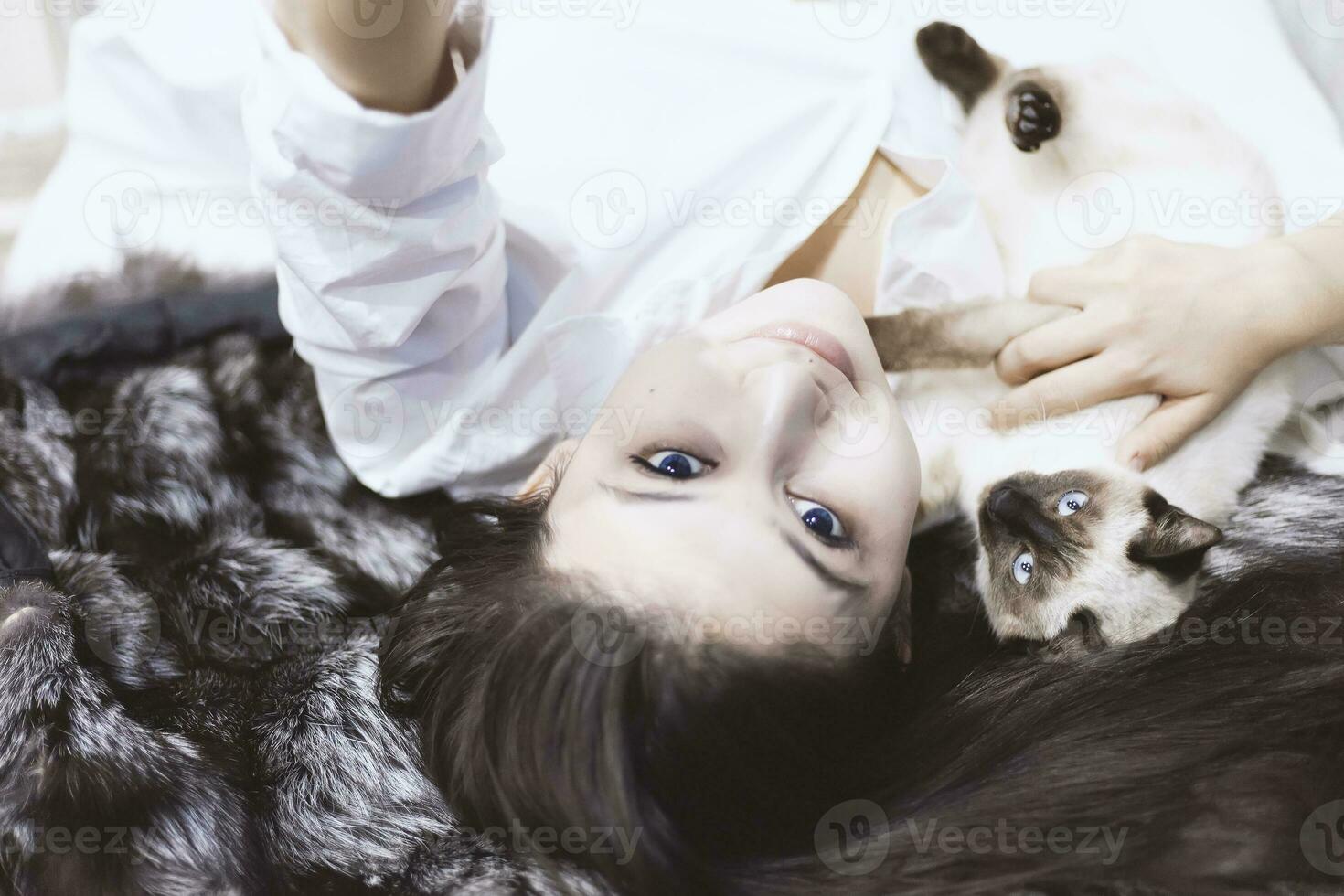 Young middle-eastern woman lying on bed and having fun with her Siamese cat. Pet loving concept. View from above to cute girl playing with cute kitten photo